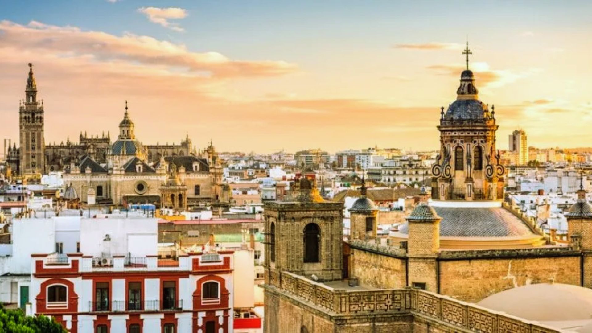 Historic cityscape of Seville at sunset, featuring ornate architectural landmarks like cathedrals and towers, under a sky with soft orange hues.