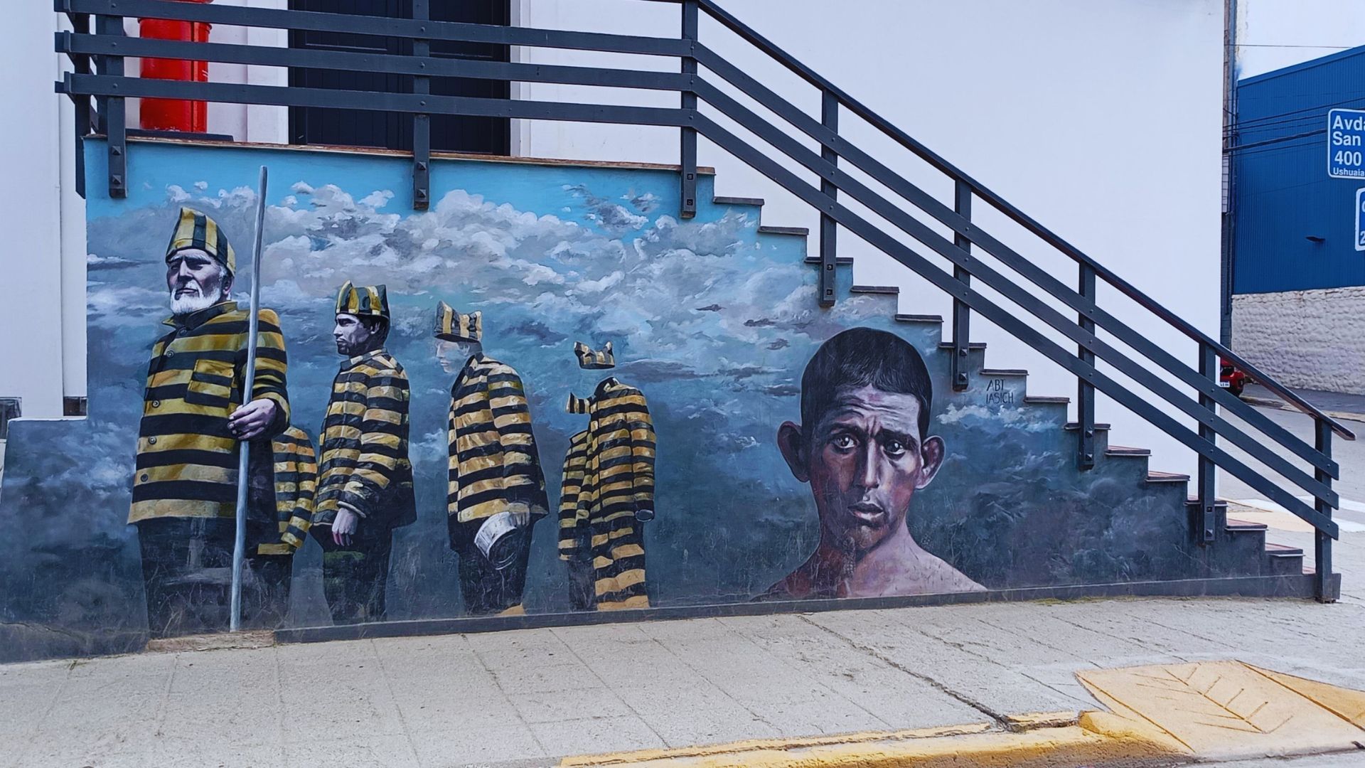 Mural on stair wall depicts four men in striped prison uniforms walking toward the horizon, with a realistic portrait of a shirtless man looking solemn.