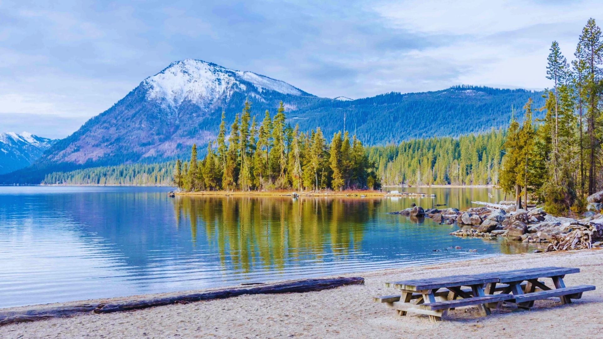 A picnic table by a lake, with mountains rising in the background under a clear blue sky.