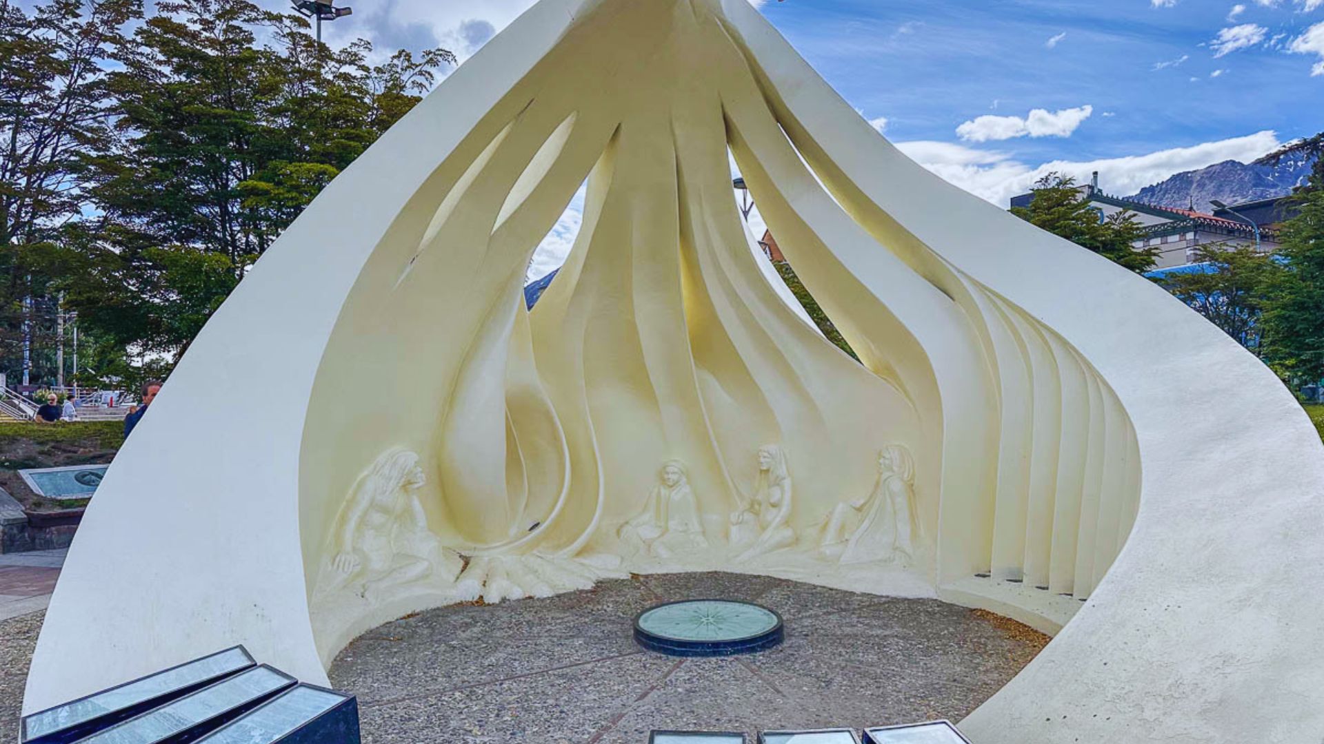 A large, curved white sculpture resembling a seashell with carvings of people inside, set outdoors against a backdrop of trees and a blue sky.