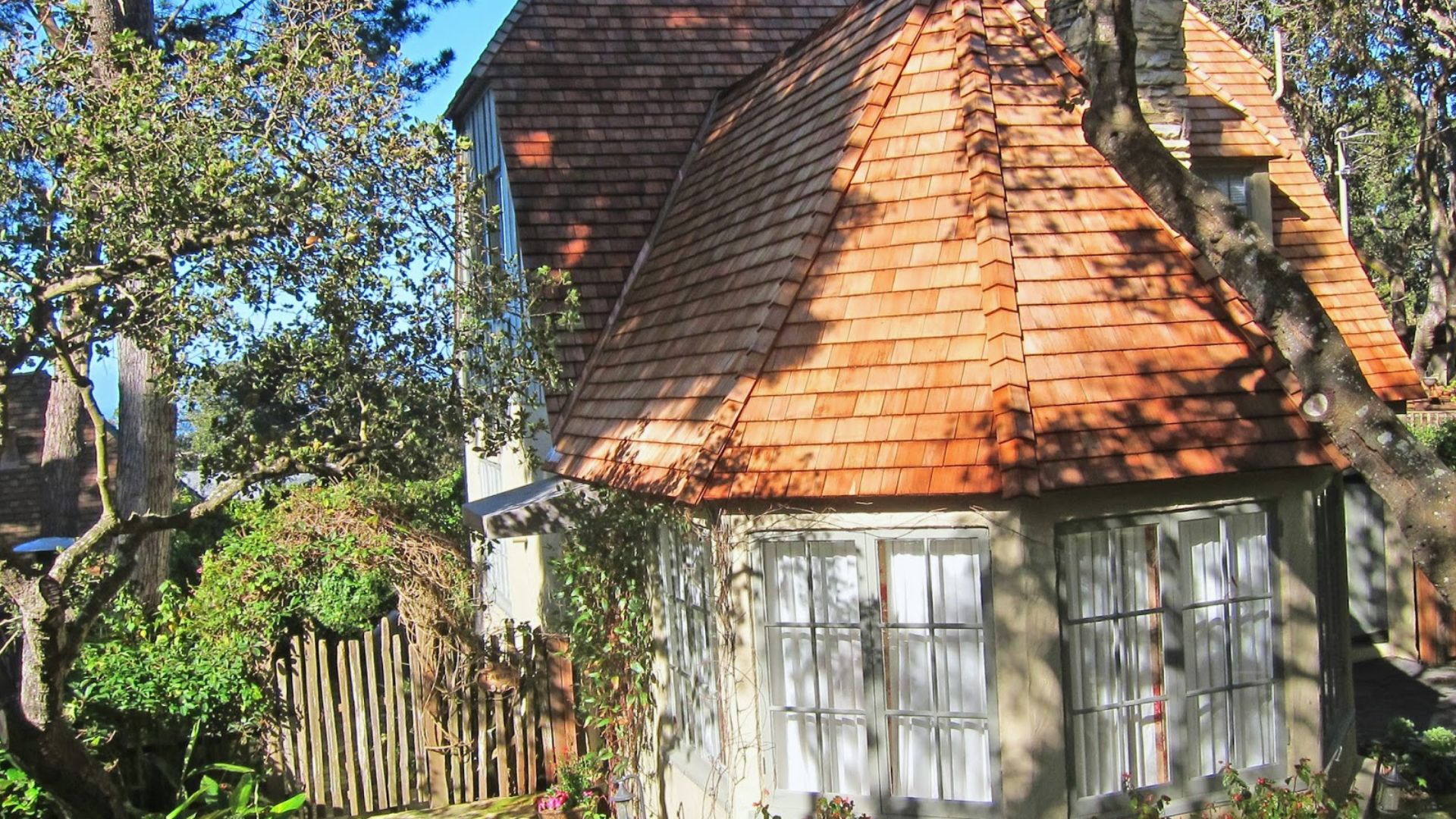 A cozy cottage with a steep wooden shingle roof and large paned windows, nestled among trees and shrubs, conveys a peaceful, rustic charm.