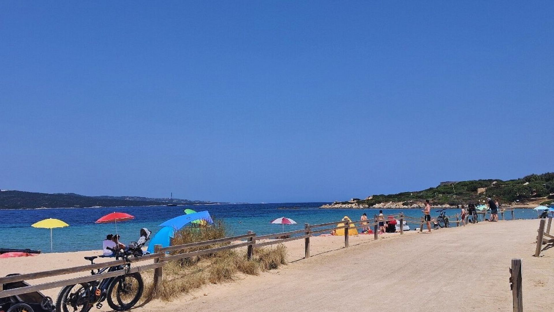 Sunny beach scene with vibrant umbrellas dotting the sand, a few bicycles parked near a fence, and people relaxing by the clear blue sea under a cloudless sky.