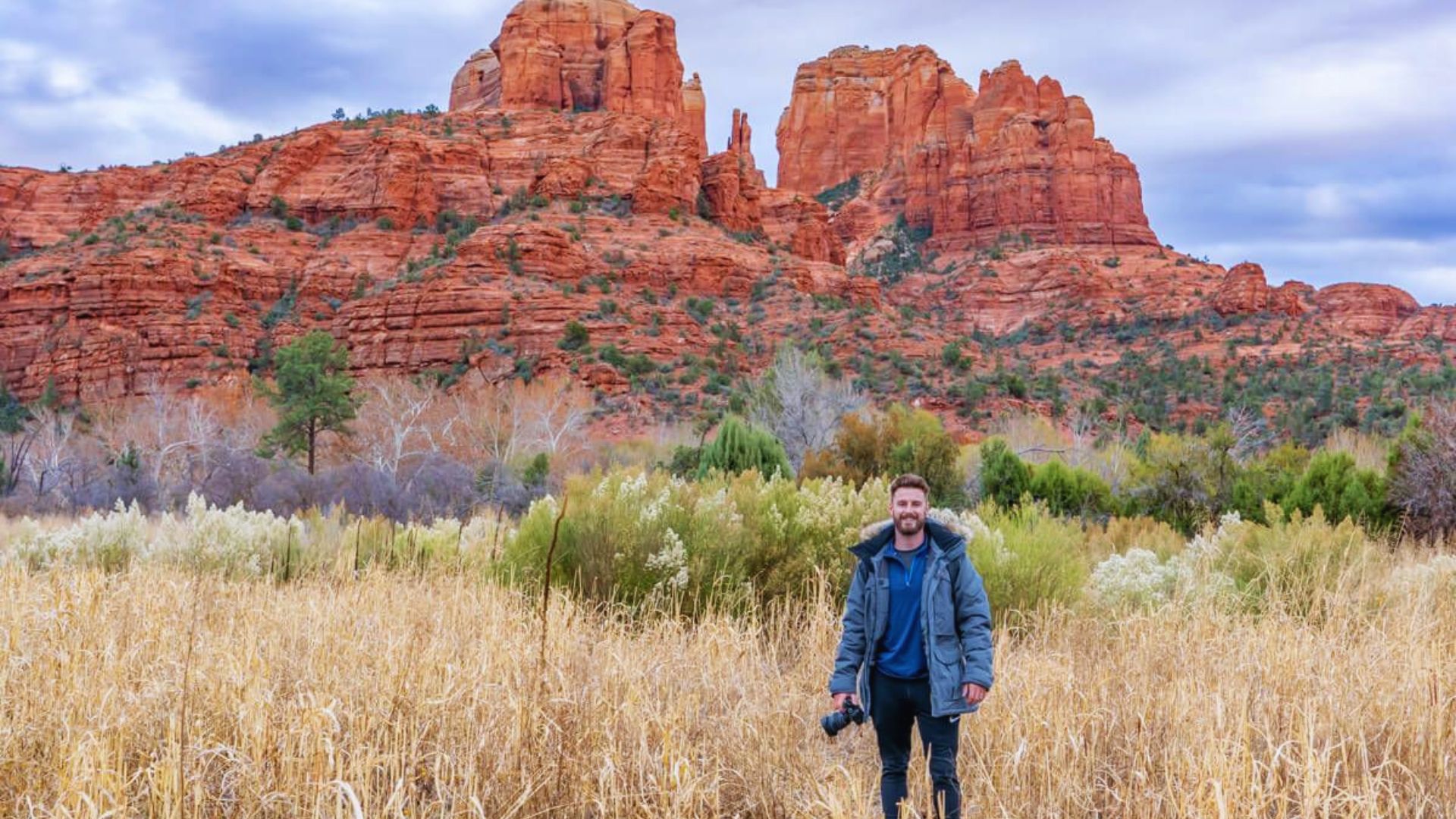 A person stands in a field of tall dry grass, holding a camera, with vibrant red rock formations in the background under a cloudy sky, conveying a sense of adventure.
