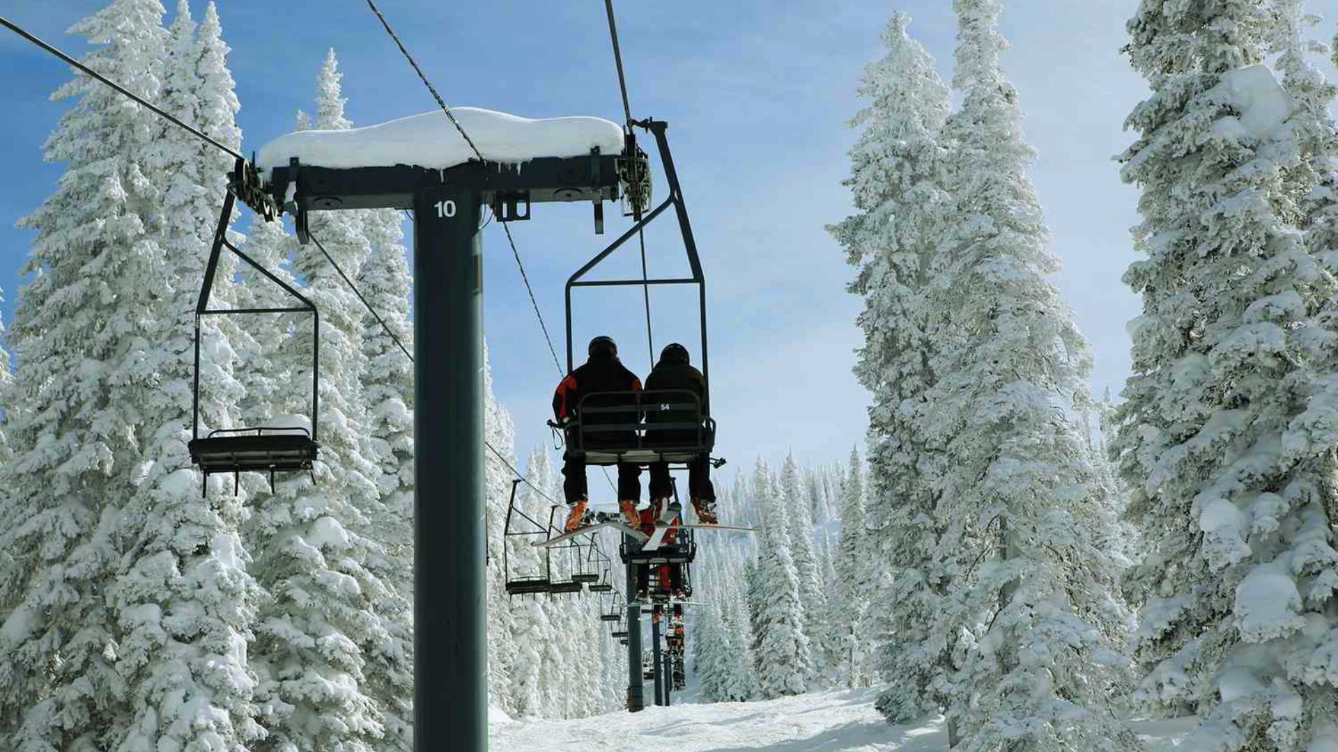 Ski lift with two people ascending a snowy mountain, surrounded by snow-covered pine trees under a clear blue sky; conveys a serene winter scene.