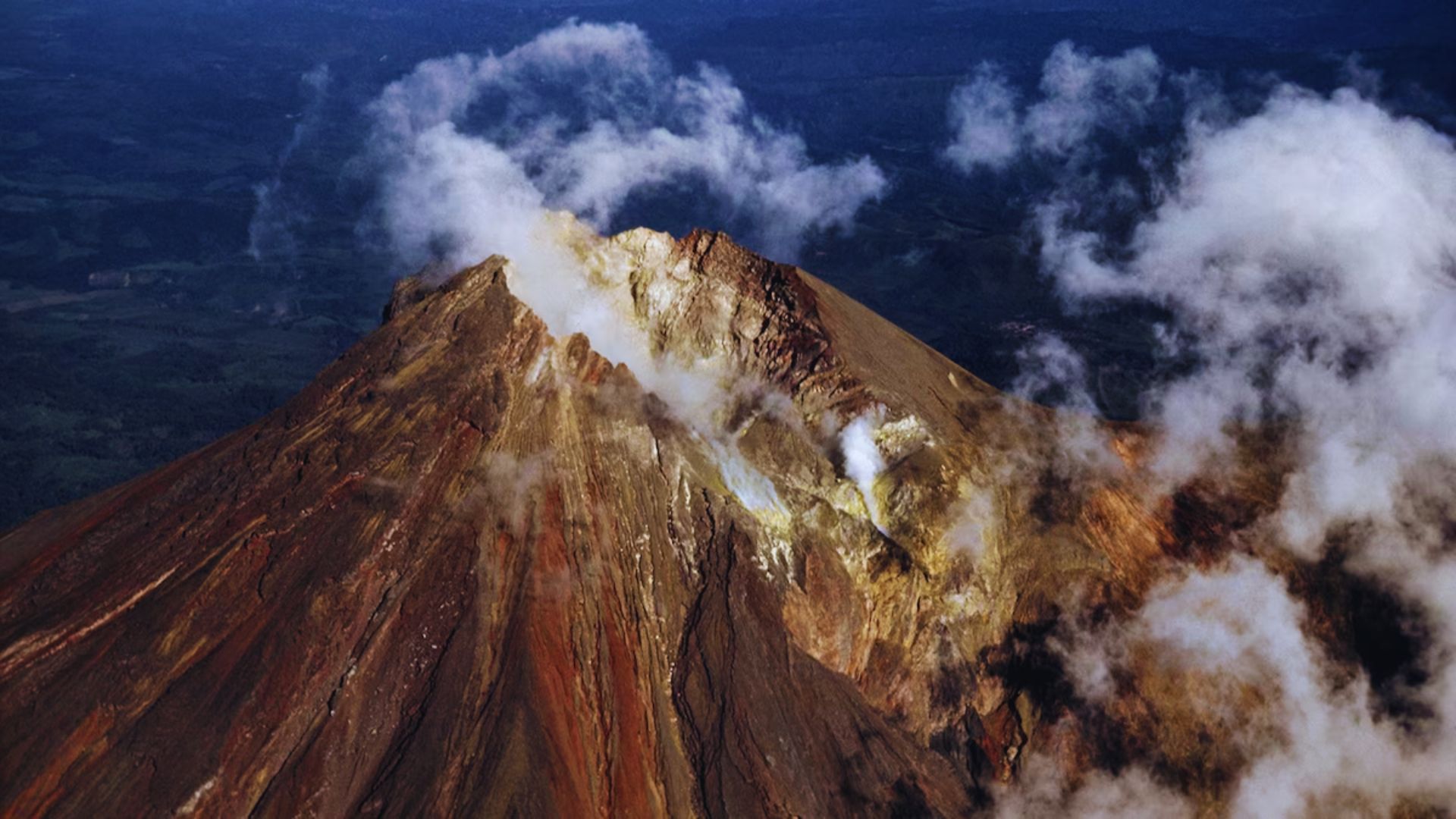 Aerial view of a volcano surrounded by clouds under a blue sky, showcasing its rugged terrain and natural beauty.