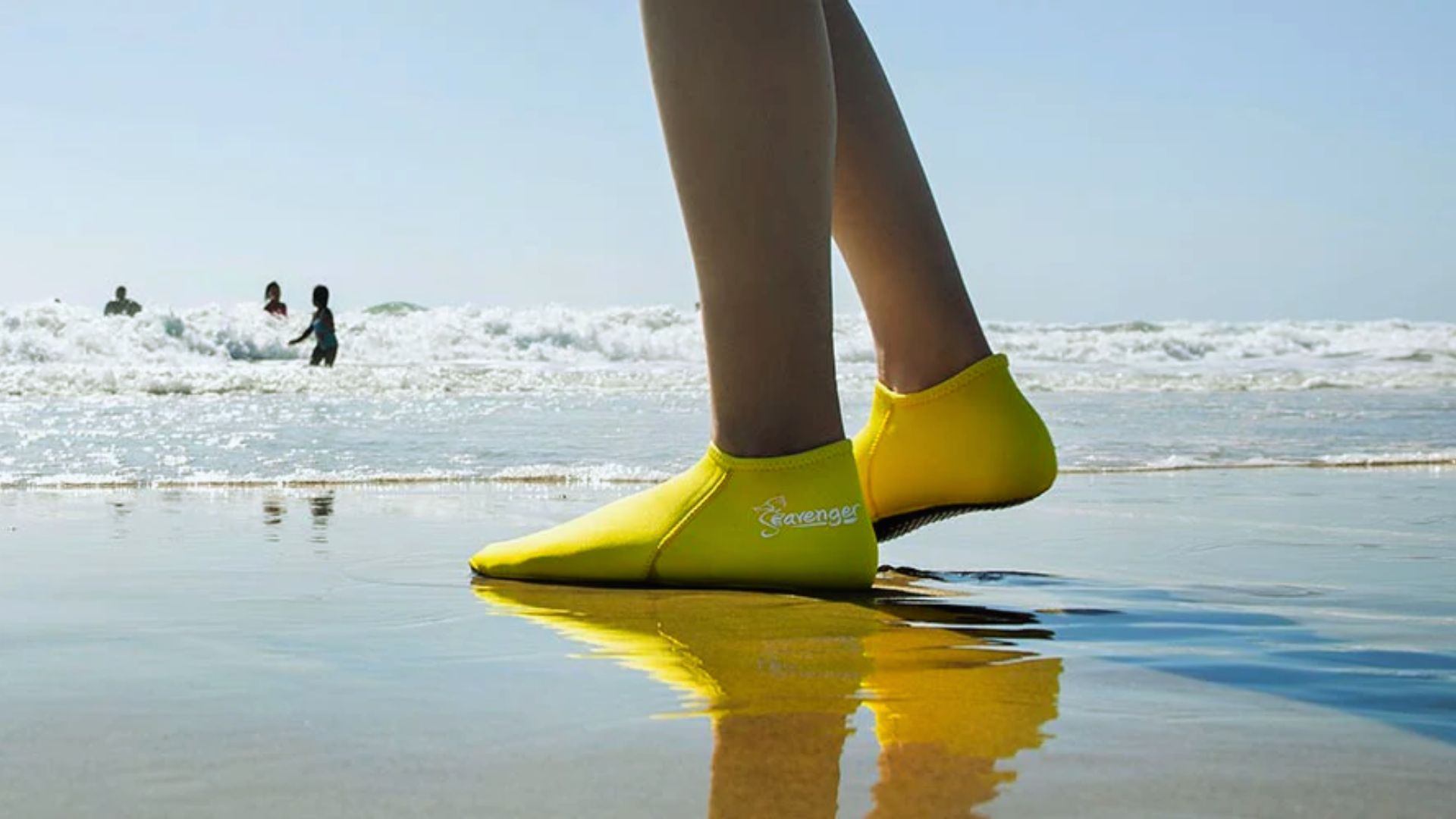A person stands on the beach, wearing bright yellow shoes, with sand and waves in the background.