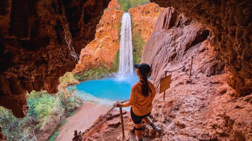 A person in an orange shirt and cap stands in a rocky cave, viewing a tall waterfall cascading into a vibrant blue pool, framed by red rock cliffs.