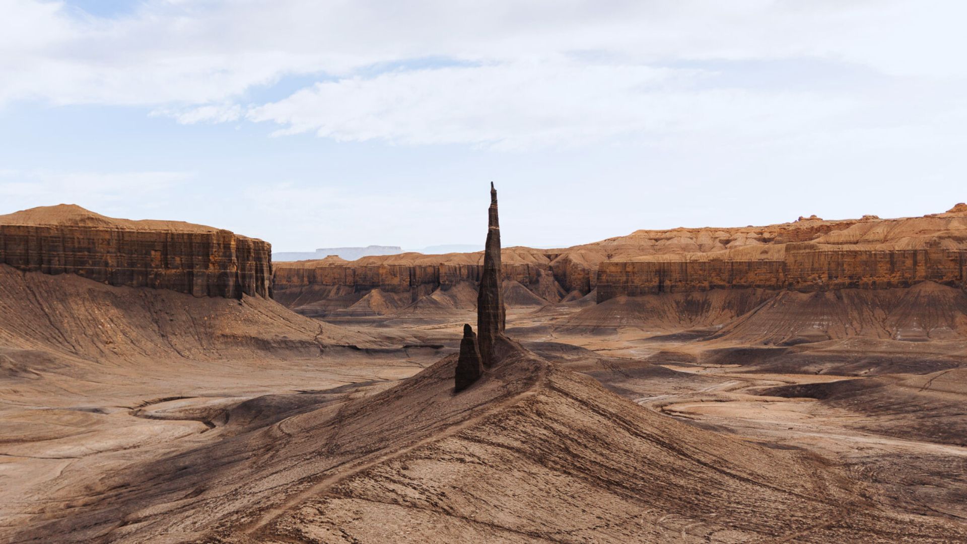 A tall, thin rock formation rises from an expansive desert landscape with layered, brown cliffs under a partly cloudy sky, conveying solitude and majesty.