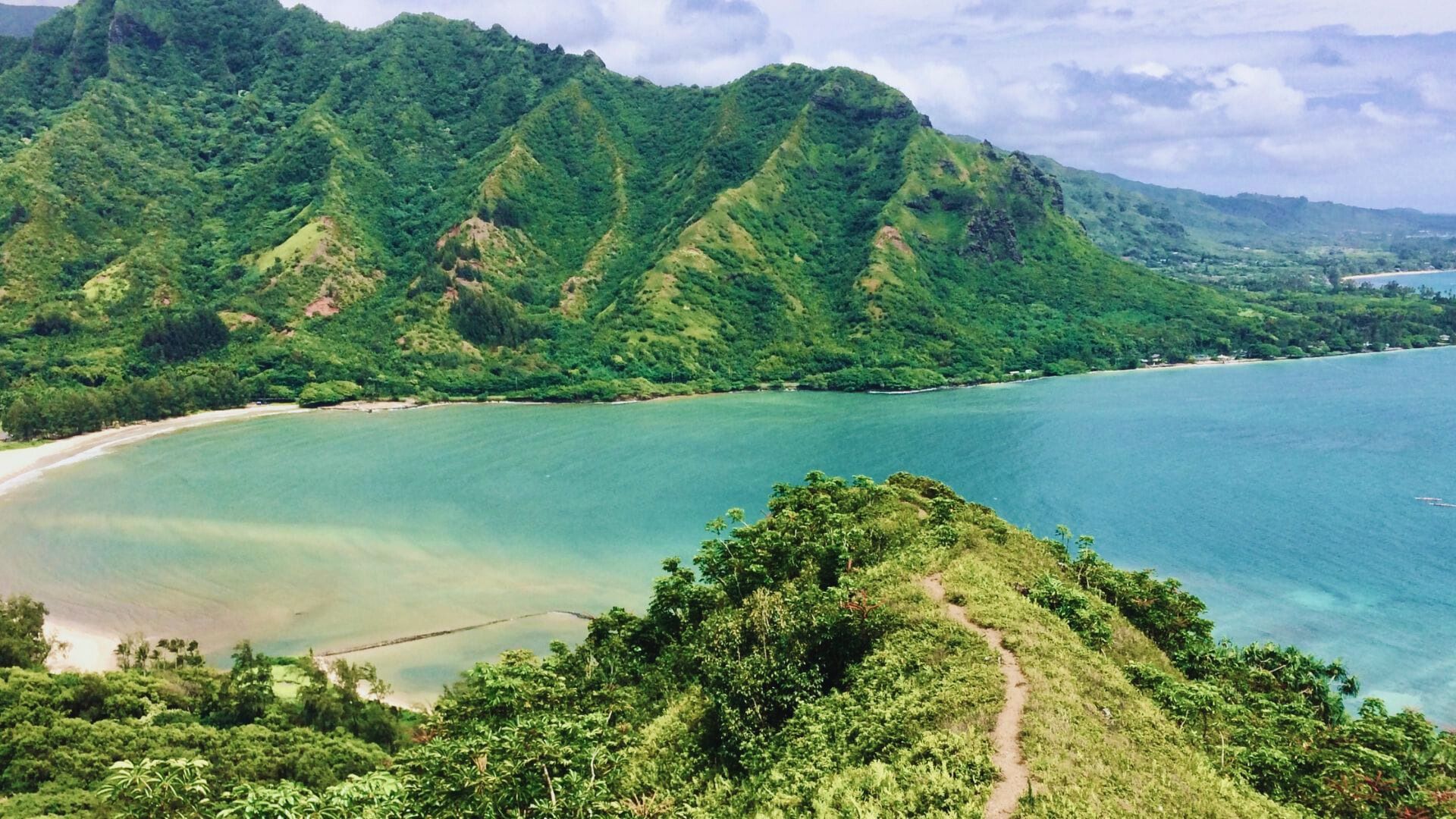 Lush green mountains overlooking a turquoise bay with a sandy shoreline. A narrow trail leads through dense foliage, evoking a sense of adventure.