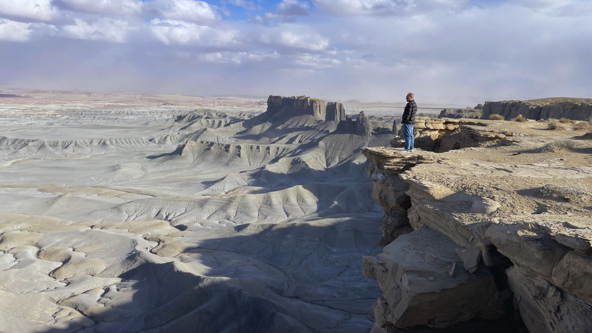 A person stands on the edge of a rugged cliff, overlooking a vast, barren desert landscape with distant plateaus under a partly cloudy sky.