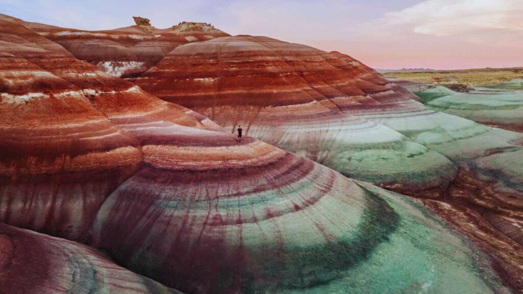 Vibrant layered hills with hues of red, pink, and green under a colorful sky. A small figure stands atop, emphasizing the vastness and beauty.