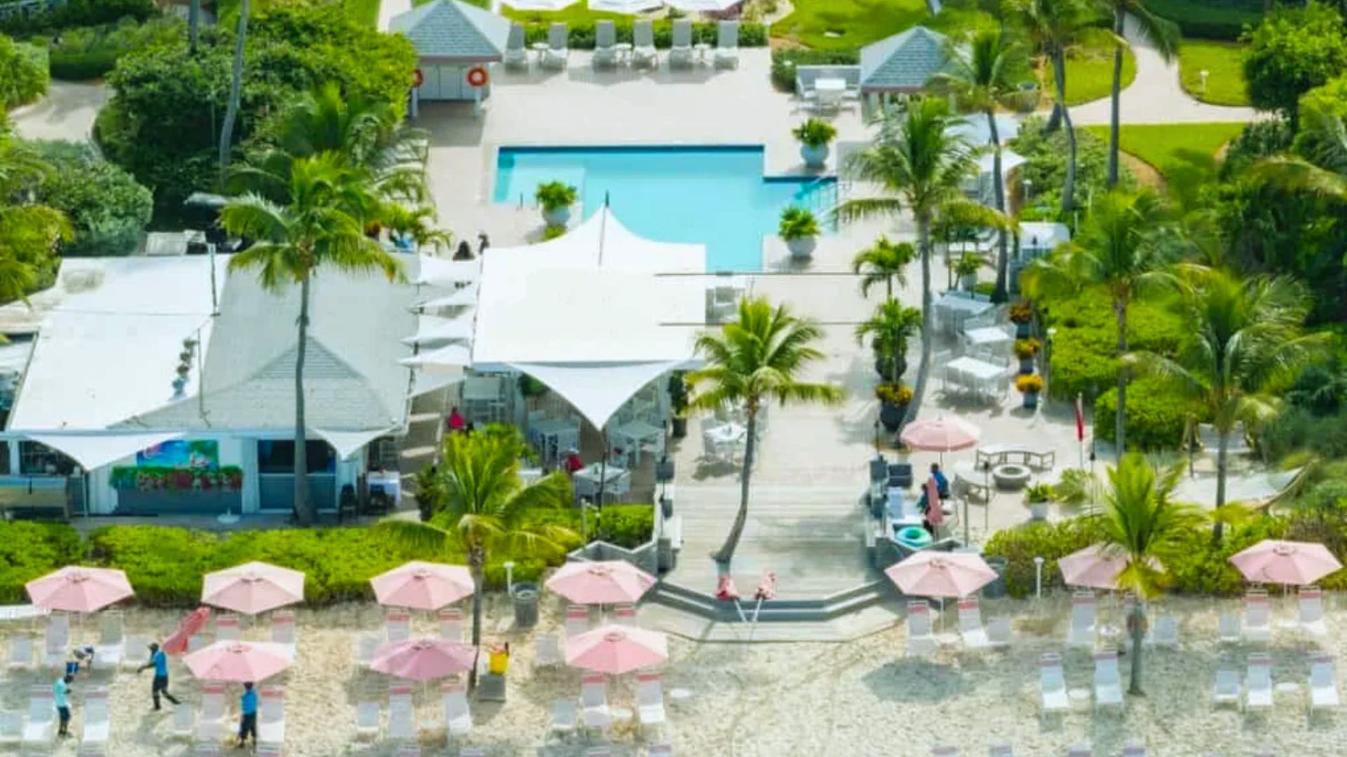 . Aerial view of a resort featuring a large pool and a sandy beach, surrounded by lush greenery and sun loungers.