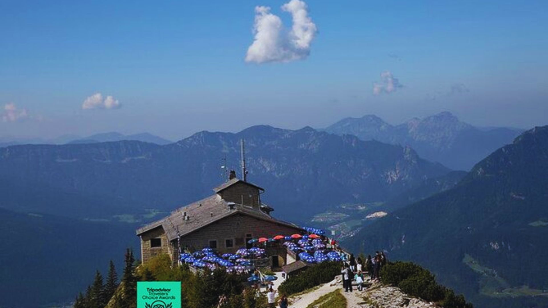 A small building atop a mountain with several people seated on the ground, enjoying the scenic view around them.
