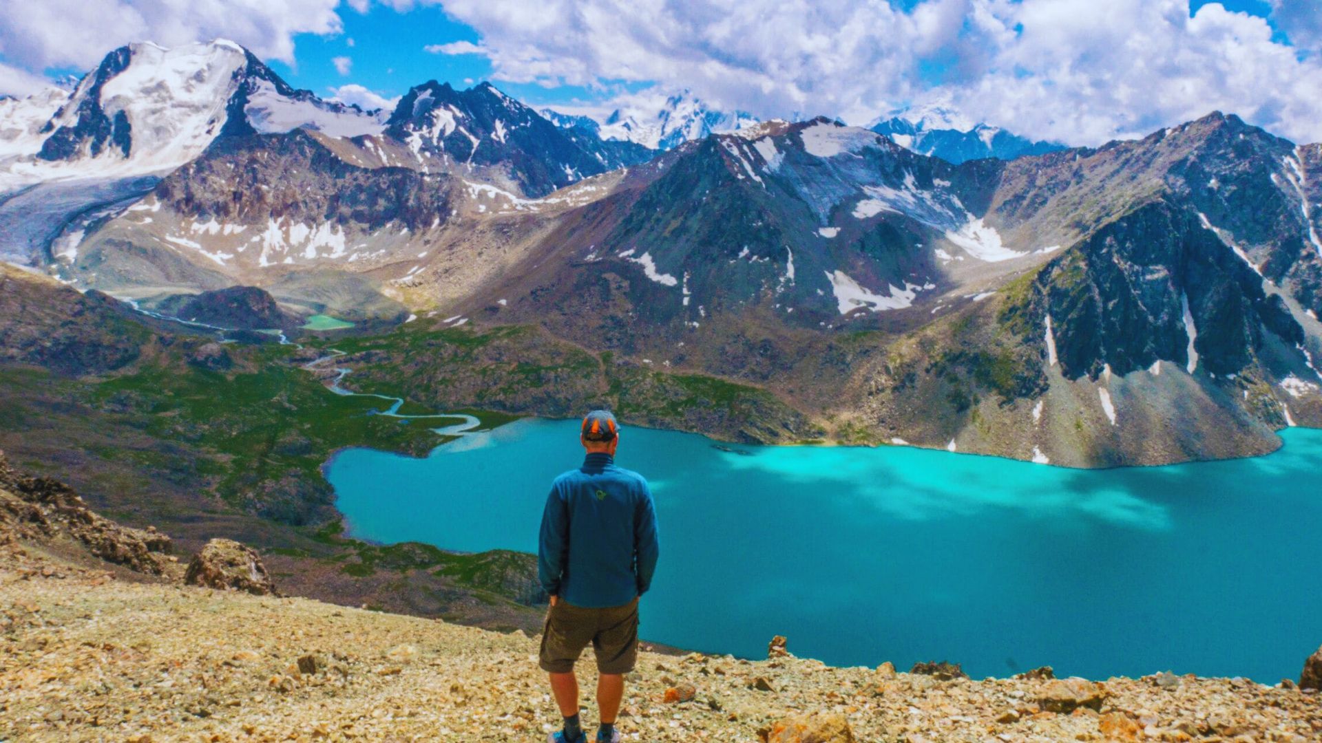 A man stands on a mountain peak, gazing at a serene lake below, surrounded by lush greenery and distant hills.