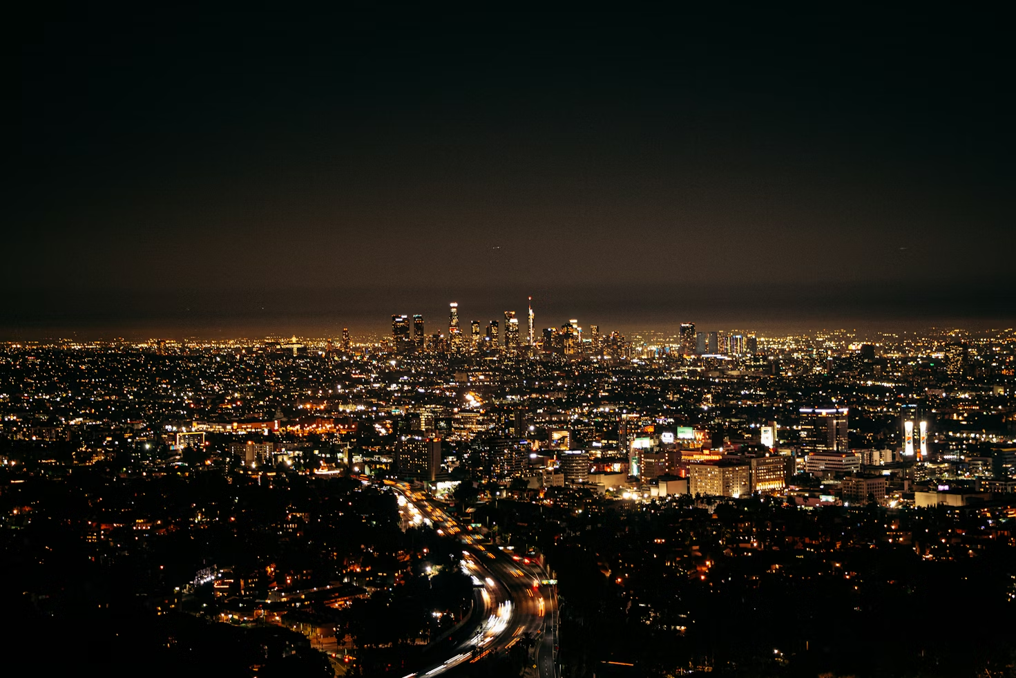 Nighttime panorama of a bustling cityscape with glowing skyscrapers in the distance. Bright car lights form streaks on winding roads, creating a lively, dynamic scene.