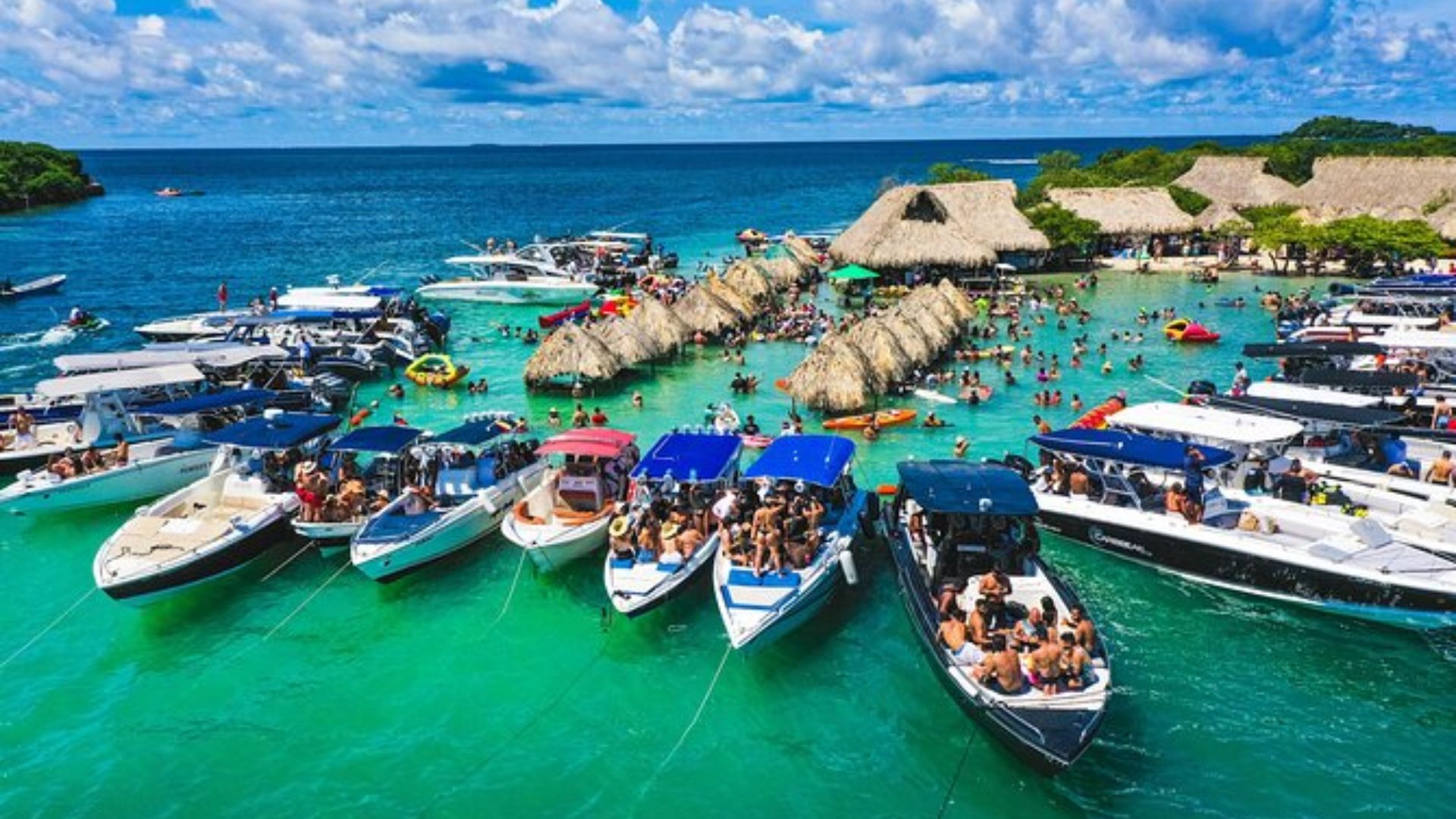 A large group of boats docked at a sandy beach, with clear blue water and a bright sky in the background.