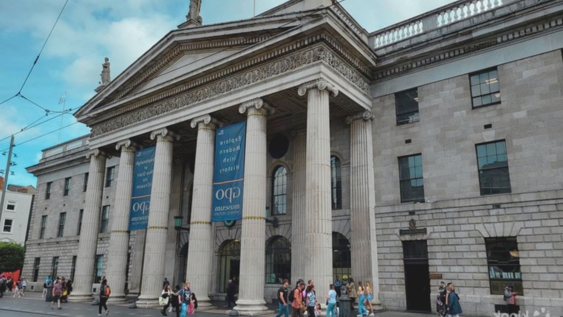 A group of people walking along the street in front of a large building on a sunny day.