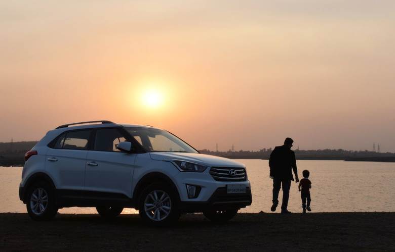 A white SUV is parked near a serene lake at sunset. In the foreground, a silhouette of an adult holding a child's hand creates a peaceful, warm scene.