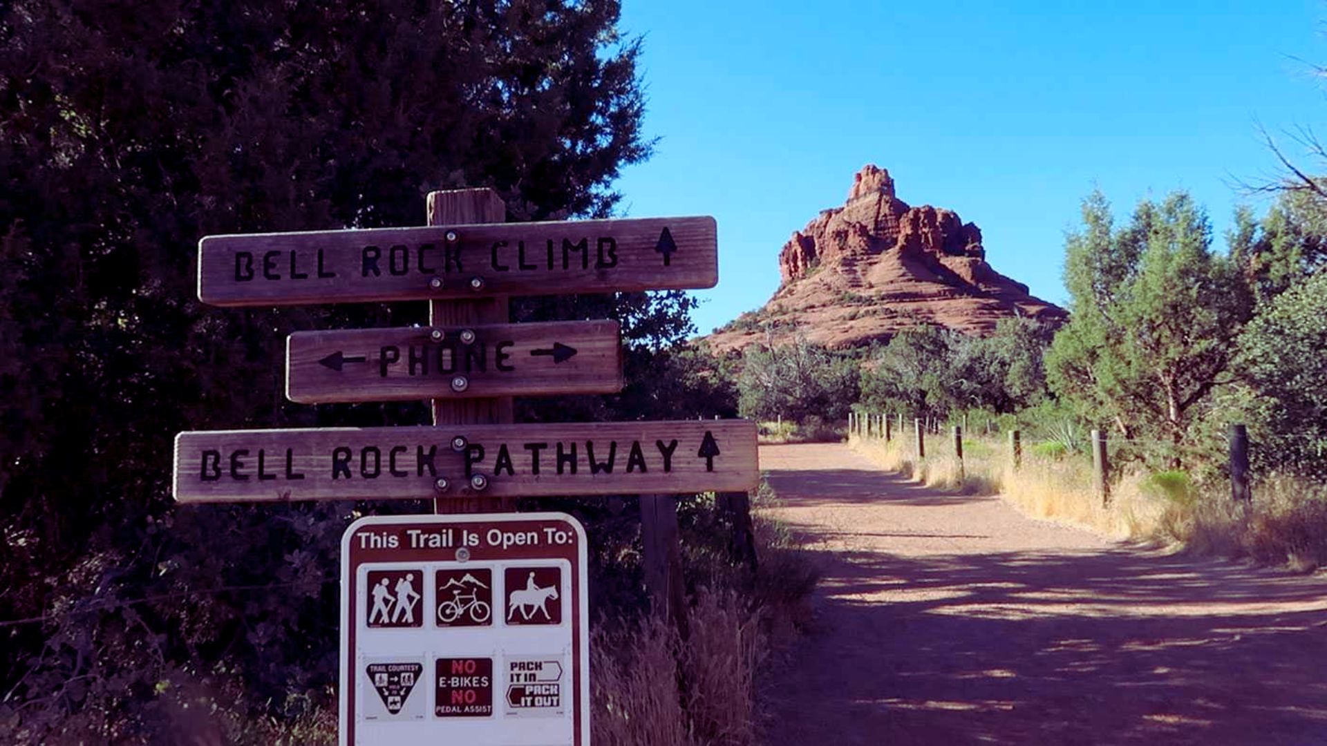 Wooden trail sign for Bell Rock Climb and Pathway, with red rock formation in the background and a dirt path lined with green shrubs in Sedona.