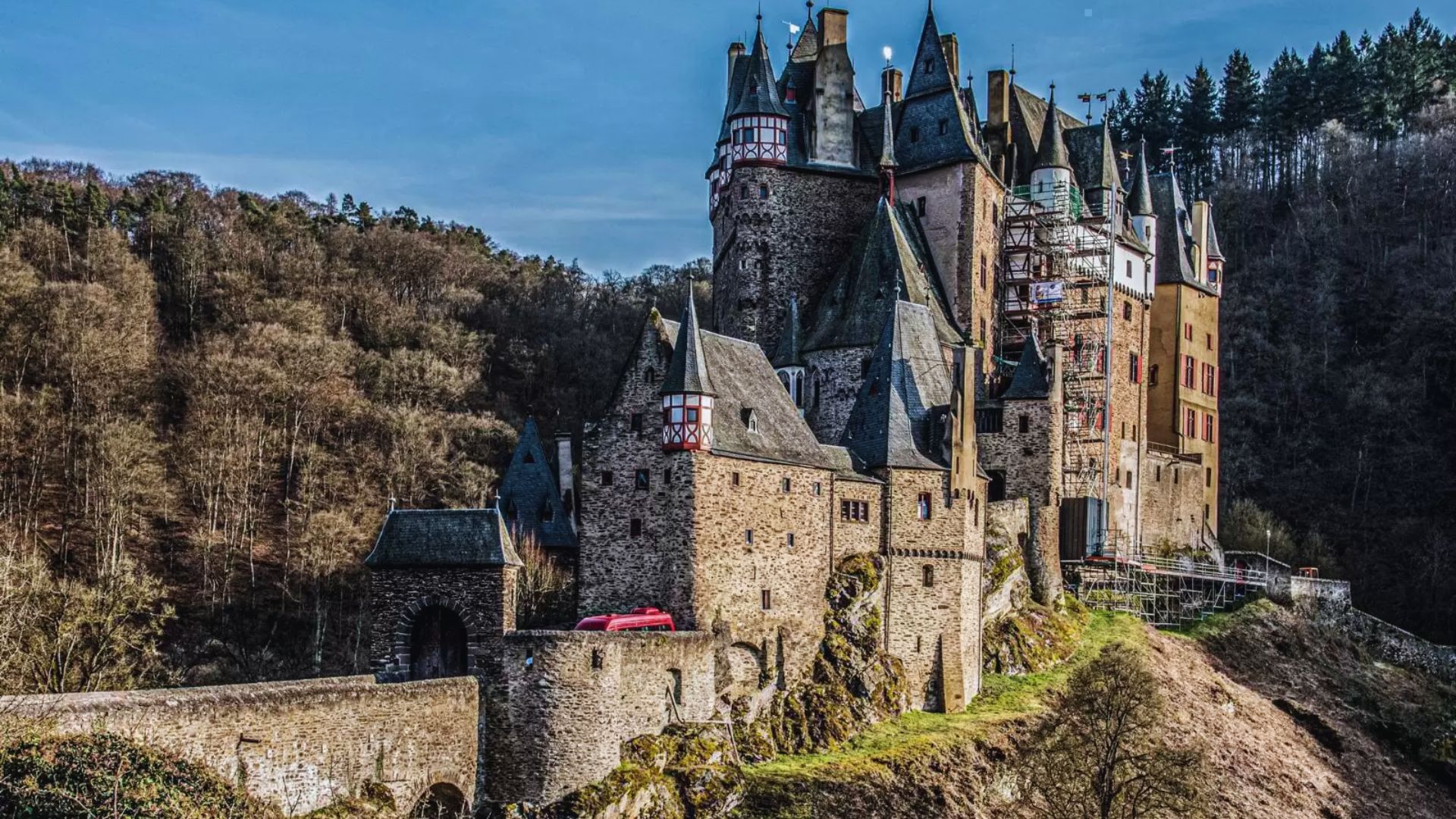 A medieval castle with tall towers and stone walls sits on a hillside surrounded by dense, leafless trees under a clear, blue sky.