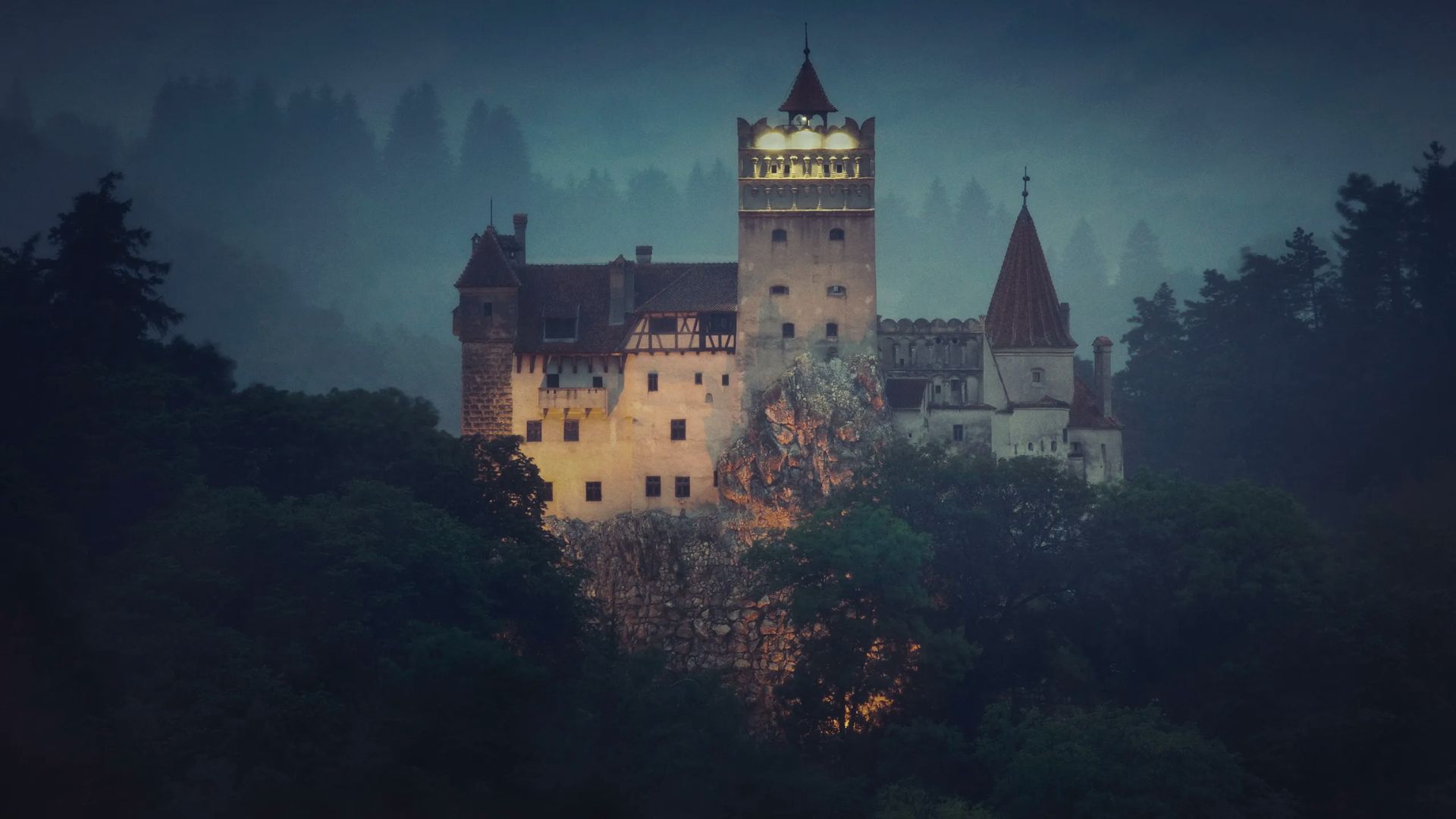 A castle with illuminated windows stands amidst a foggy landscape and dense trees, evoking a mysterious and atmospheric mood.