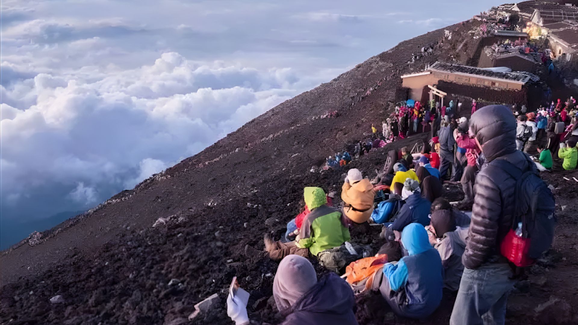Crowd sitting on a rocky mountain slope, bundled in colorful jackets, watching a sunrise above a sea of clouds, conveying a serene, communal atmosphere.