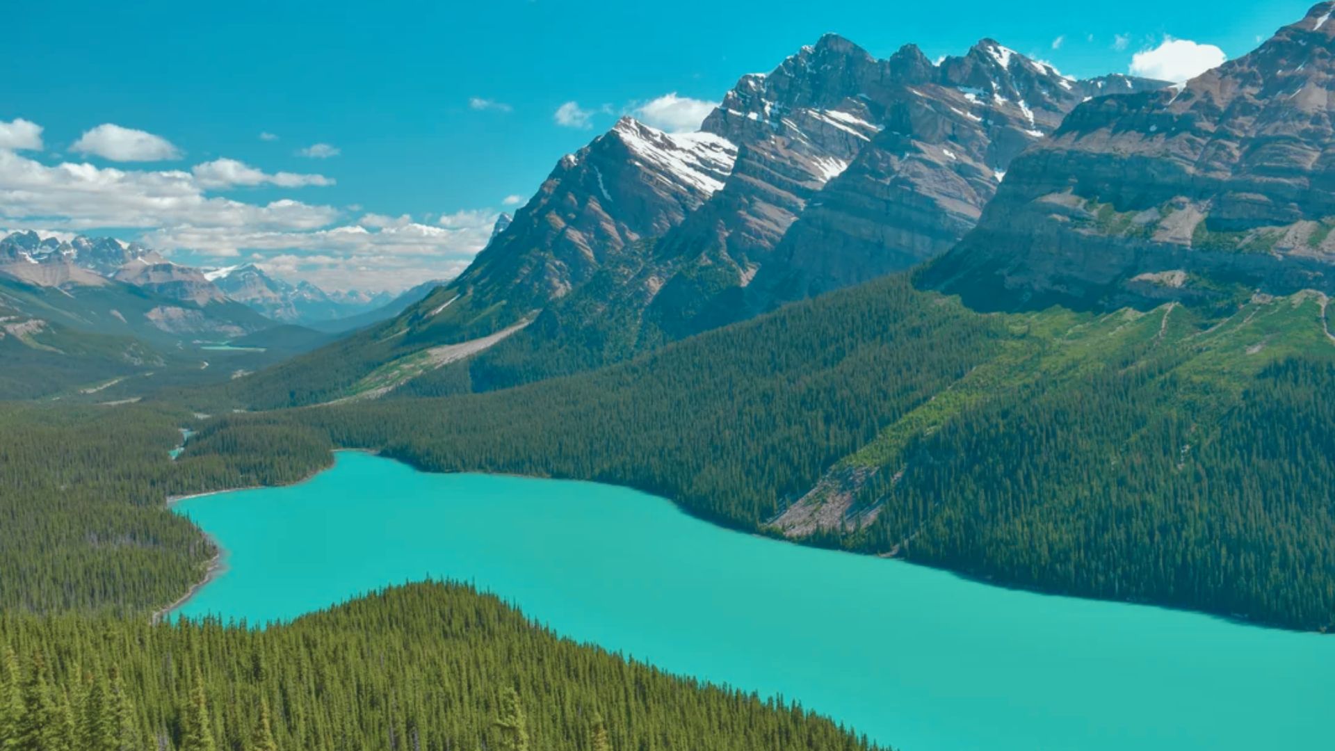 A turquoise lake winds through dense green forests, surrounded by snow-capped mountains under a clear blue sky. The scene is serene and majestic.