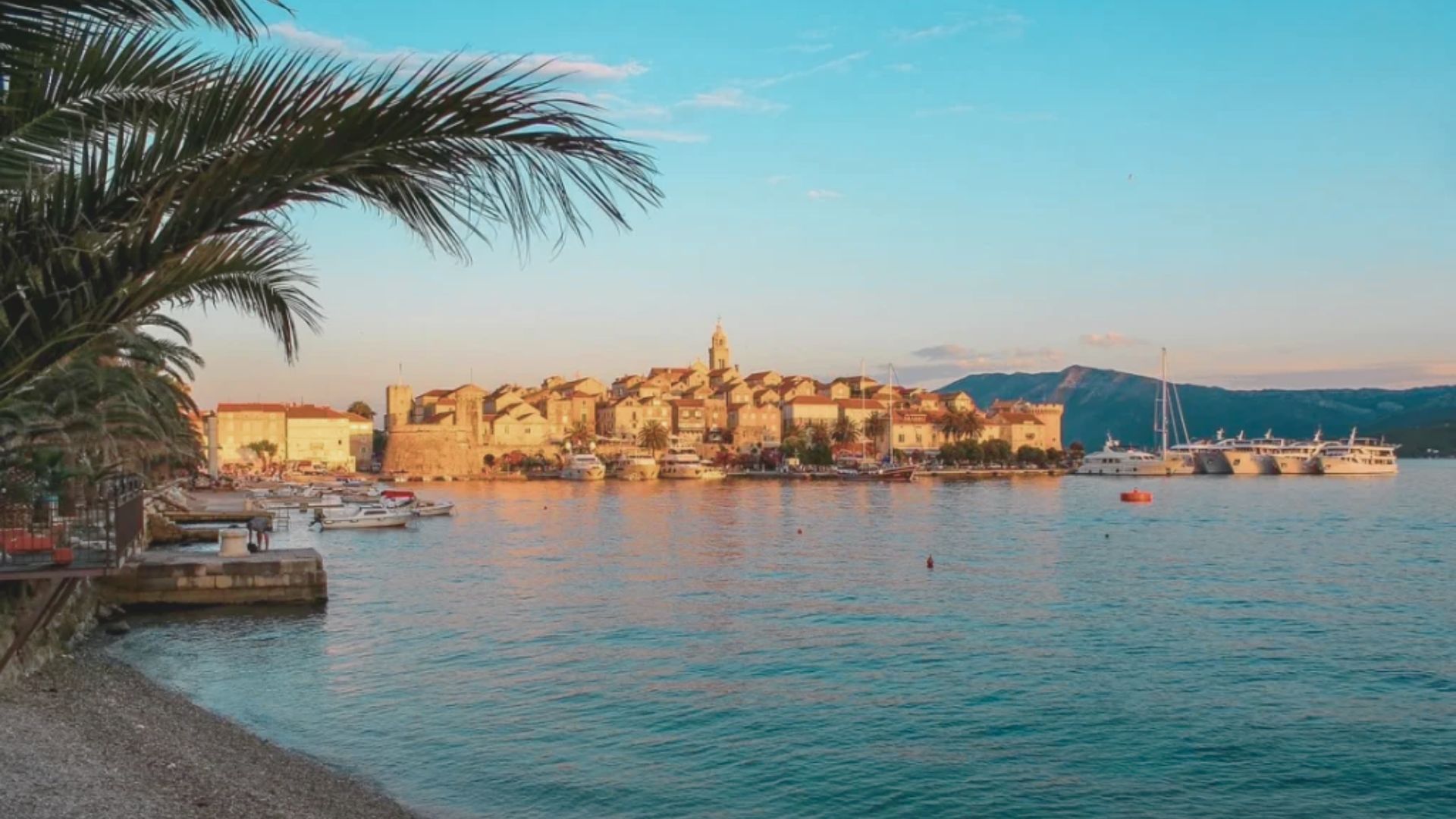Coastal town at sunset with calm water, palm trees, and boats. Warm light highlights historic buildings, creating a serene and picturesque atmosphere.