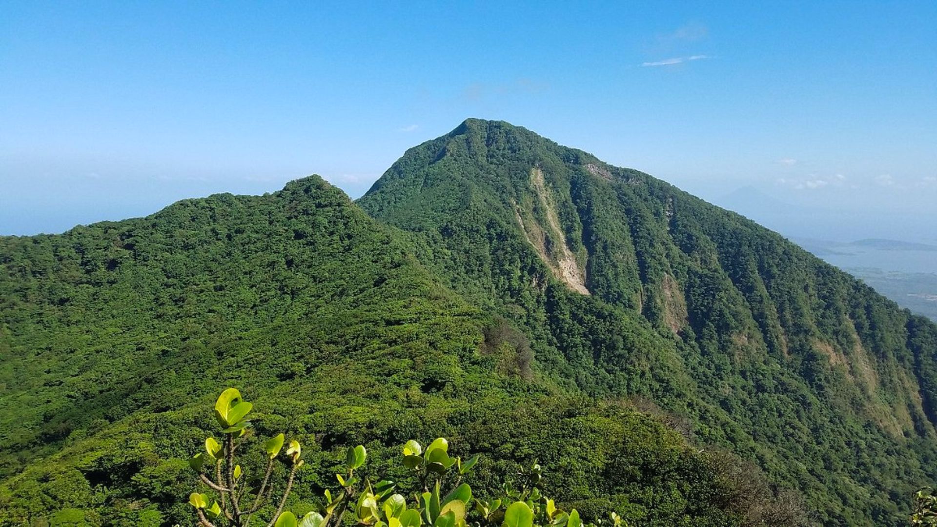 Lush green mountains under a clear blue sky, with dense foliage covering the landscape. The scene conveys tranquility and natural beauty.
