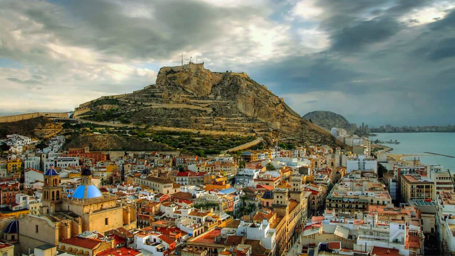 A scenic view of a coastal city with colorful buildings under a dramatic cloudy sky. A rocky hill with a historic fort overlooks the vibrant urban landscape.