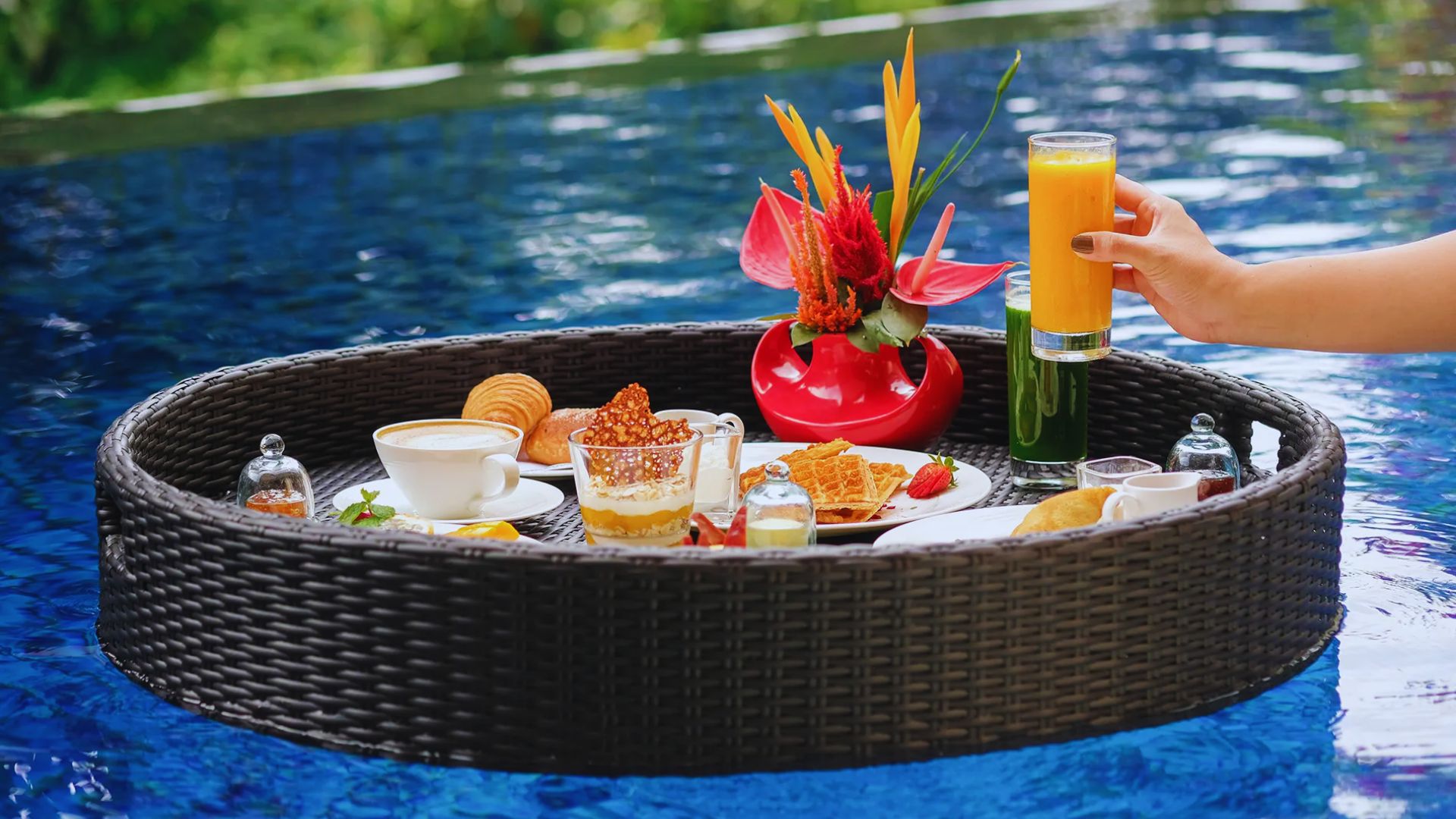 A round wicker tray floats in a pool, holding a colorful breakfast with waffles, pastries, tropical flowers, and juice. A hand reaches for a glass.