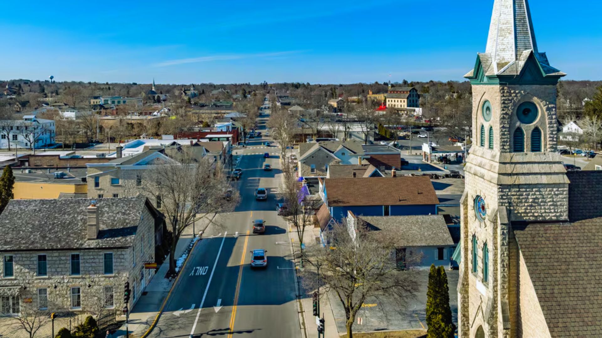 Aerial view of a small town on a clear day, featuring a large stone church with a tall steeple in the foreground. Tree-lined streets and buildings stretch toward the horizon. The scene conveys a peaceful, quaint atmosphere.