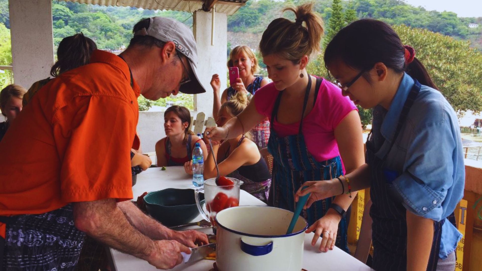 A cooking class on a porch overlooking greenery; an instructor in orange assists two women making a dish. Others watch, capturing the moment.
