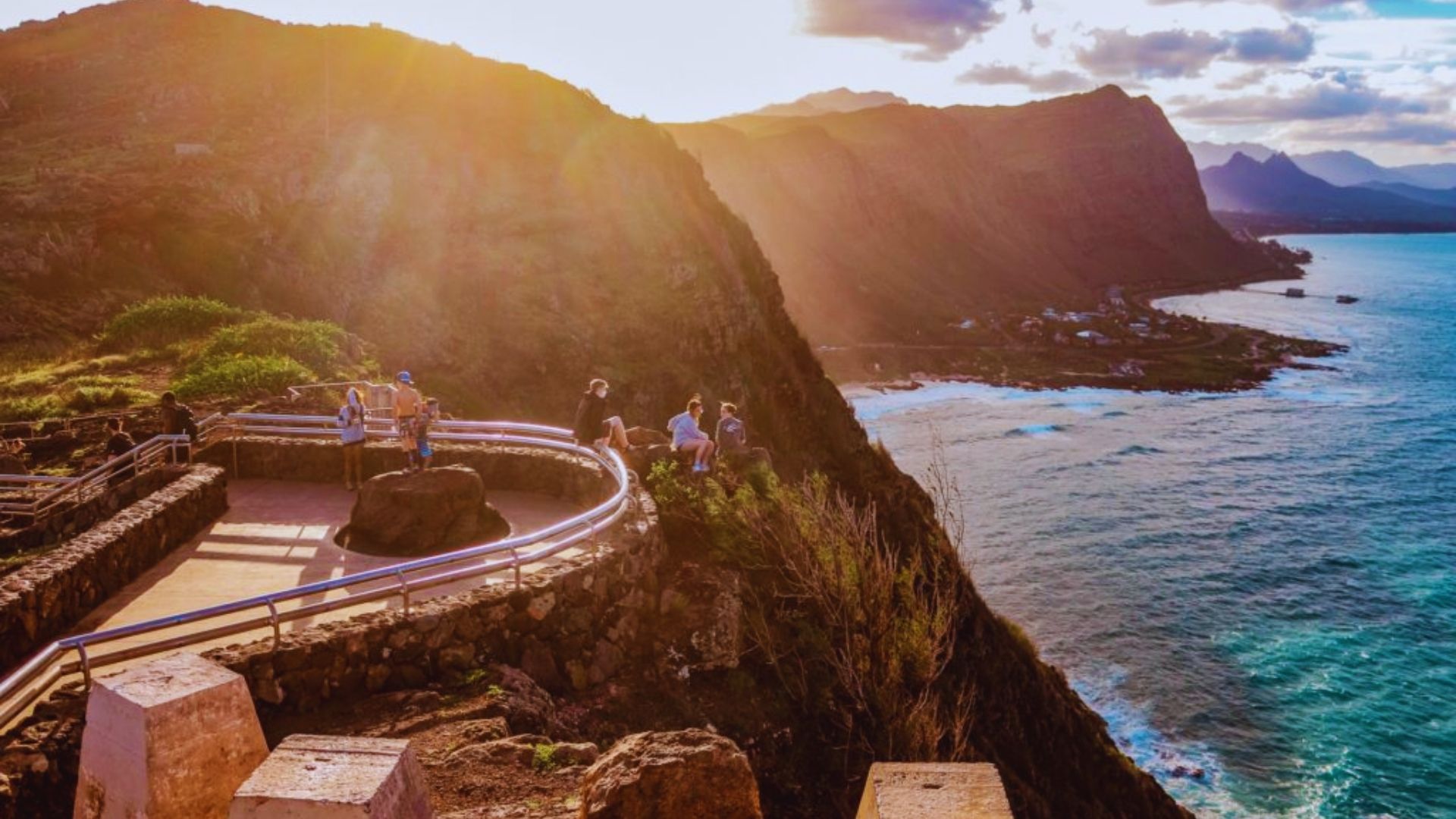 Scenic overlook with visitors enjoying a stunning sunset view of ocean cliffs and mountains. Warm light creates a serene, inspiring atmosphere.