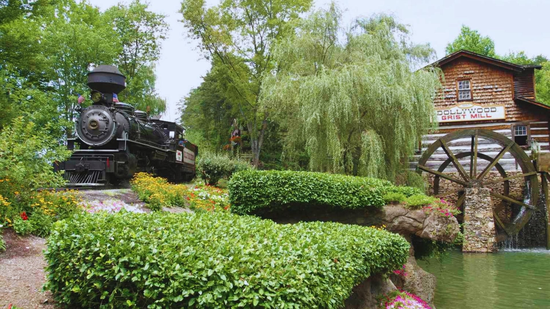 A vintage steam train passes lush greenery and colorful flowers near a wooden watermill labeled "Dollywood Grist Mill," evoking a nostalgic and serene atmosphere.