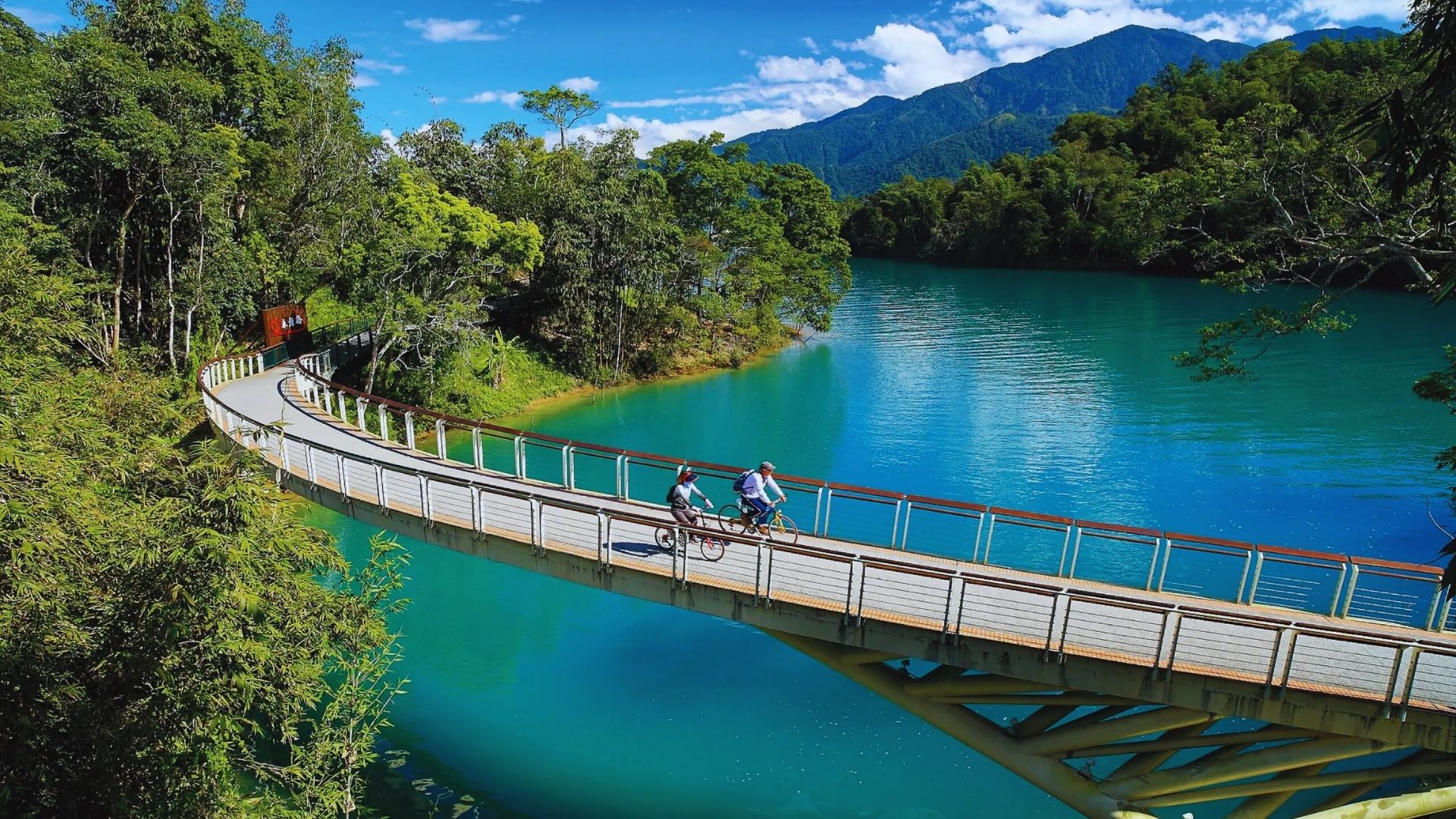 Two cyclists ride on a curved wooden bridge over a serene turquoise lake, surrounded by lush greenery and distant mountains under a blue sky.