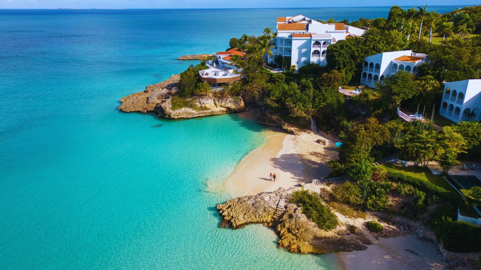 Aerial view of a tropical beach with clear turquoise water, white buildings on a lush green cliff, and a small sandy cove. Bright and serene atmosphere.