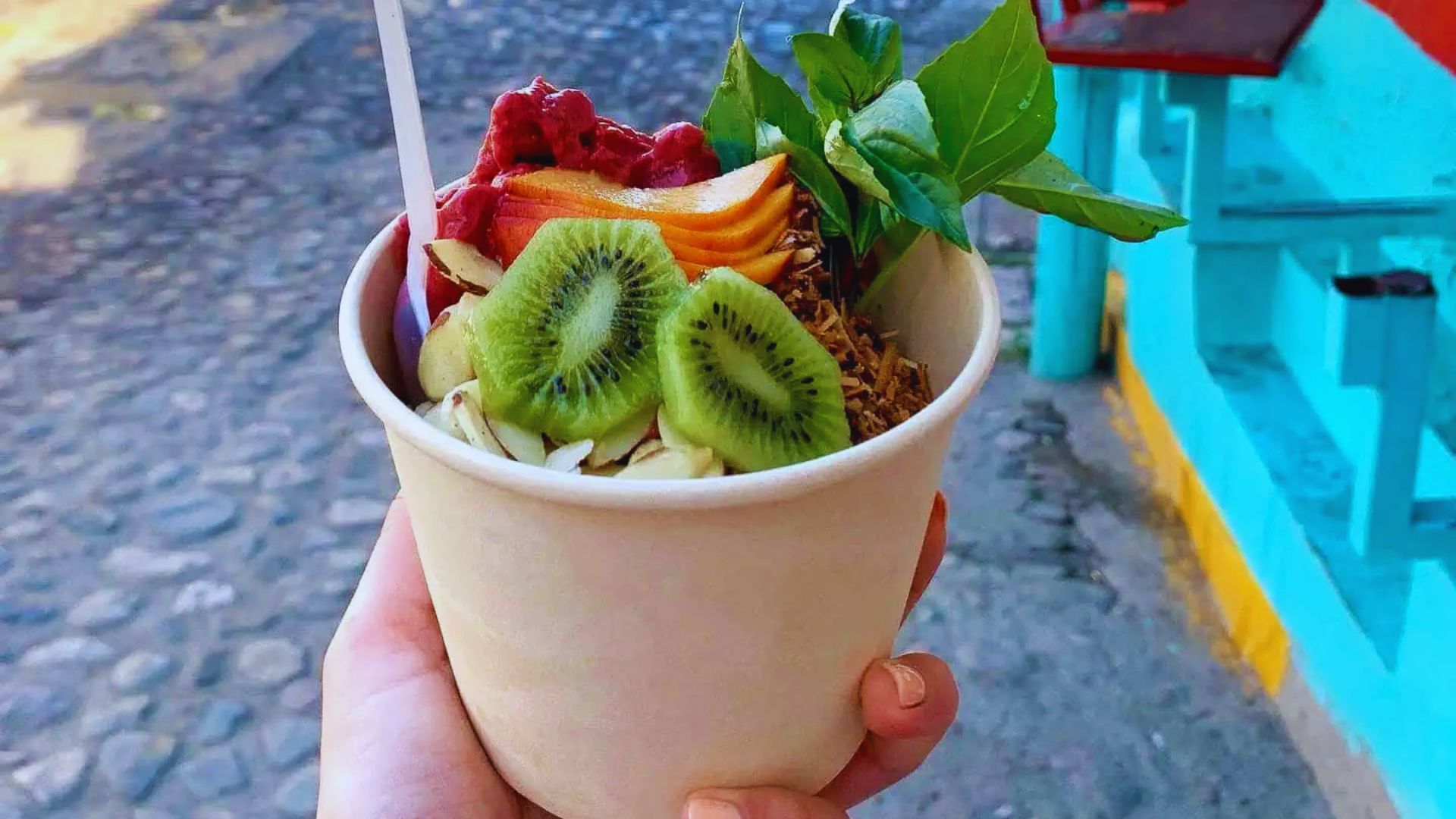 A hand holds a paper bowl filled with vibrant fruits: kiwi slices, persimmon, berries, and a sprinkle of granola. A colorful street forms the lively backdrop.