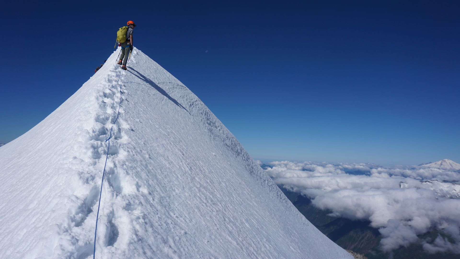 A climber wearing a green backpack and orange helmet ascends a snow-covered peak, leaving footprints. The scene conveys adventure and solitude under a clear blue sky.