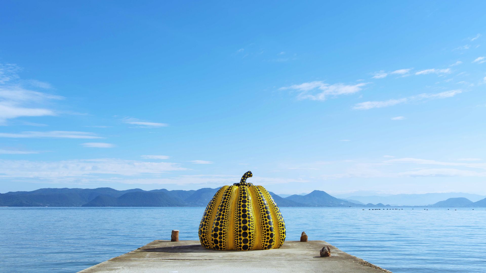 A bright yellow pumpkin rests on a wooden dock overlooking the ocean, with gentle waves lapping at the shore.