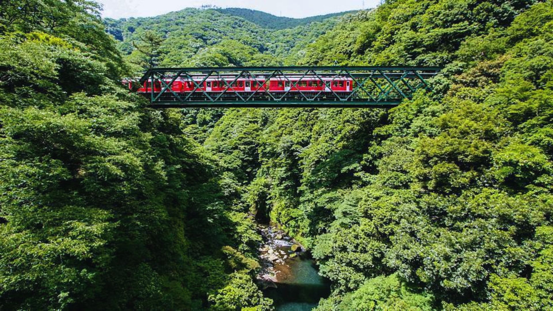 A red train crosses a green metal bridge over a lush, green forested ravine with a stream below, conveying a sense of adventure and tranquility.