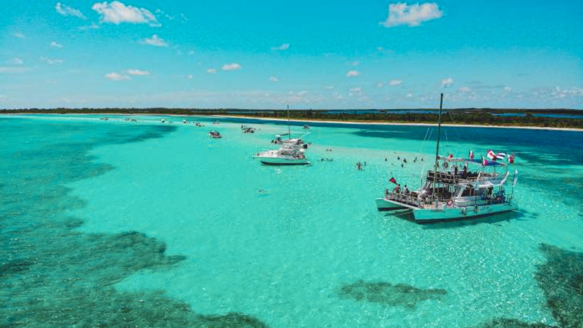 Turquoise waters with anchored catamarans and small boats. People swim nearby under a sunny blue sky. The scene feels tranquil and inviting.