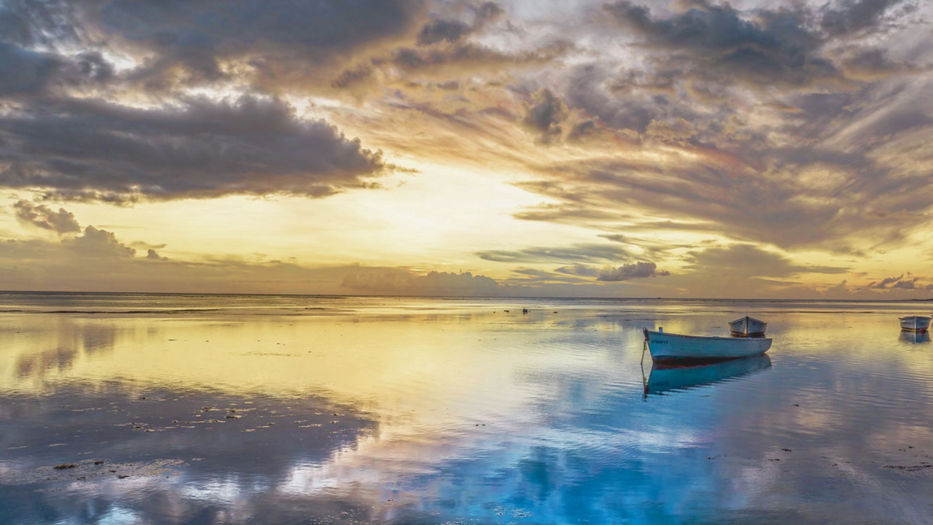 A tranquil scene at sunset with a lone boat floating on calm, reflective water. The sky is filled with dramatic clouds and warm golden hues, creating a serene atmosphere.