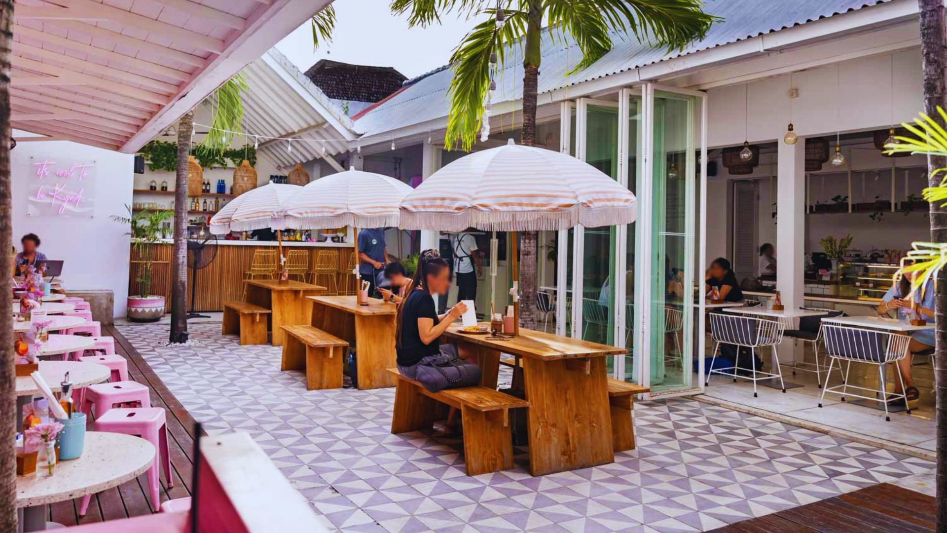 Outdoor café with wooden benches and tables, pink chairs, and striped umbrellas. Patrons are seated, enjoying a sunny day. Tropical, relaxed vibe.