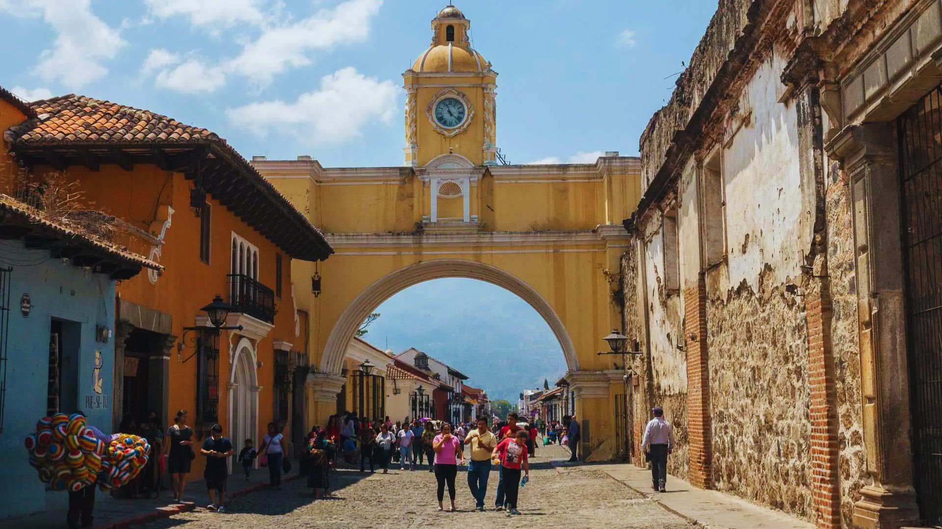 A vibrant street scene in Antigua, Guatemala, features a yellow arch with a clock. People walk along the cobblestone street, framed by colorful colonial buildings.
