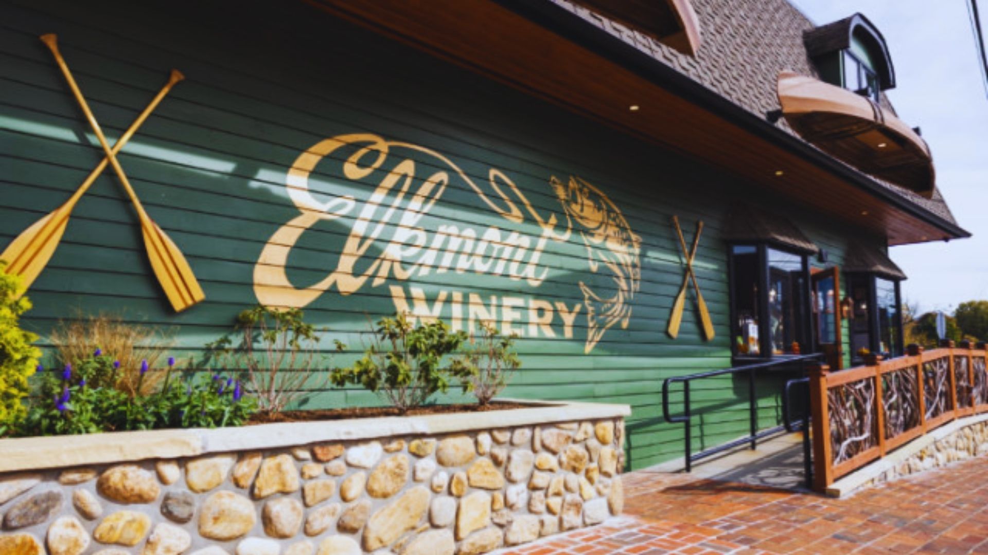Alt text: "Rustic winery entrance with green wooden facade, featuring 'Elkmont Winery' signage in cursive, flanked by paddle decor. Stone and wood details lend a warm, welcoming atmosphere."