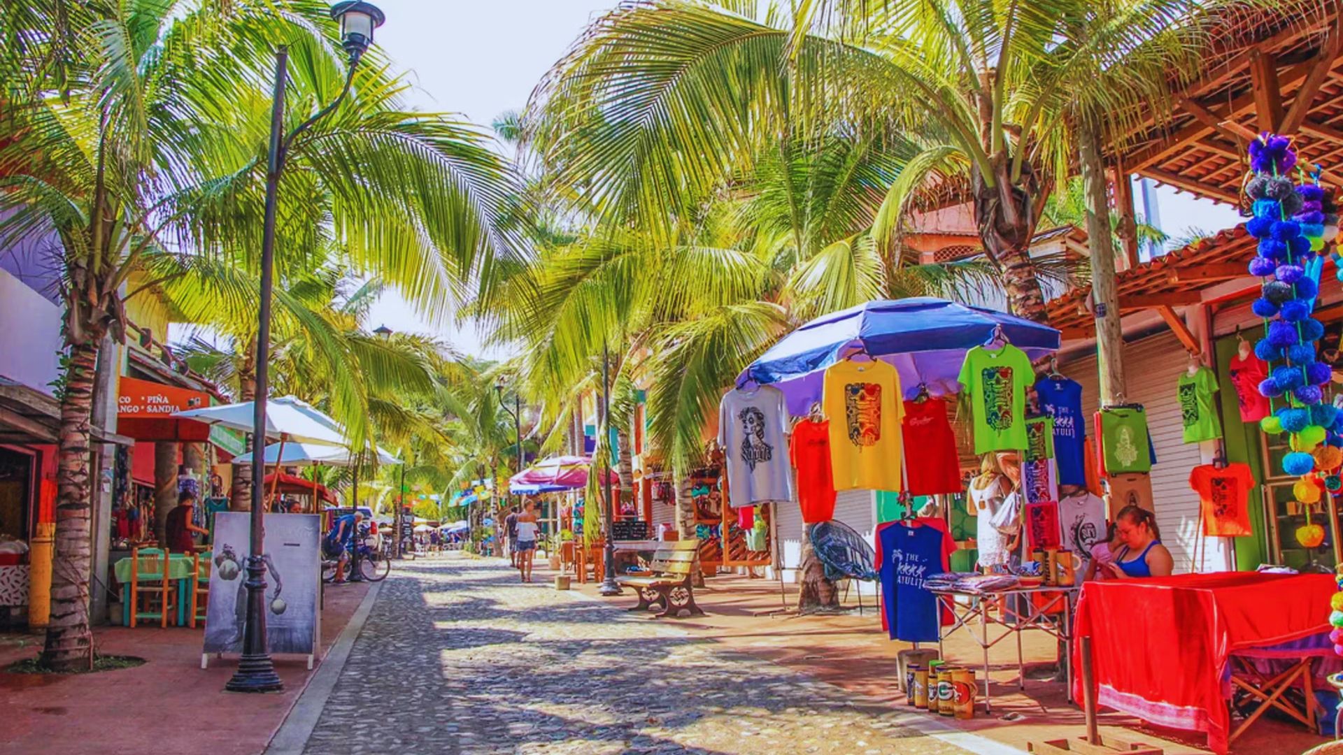 A vibrant street scene under sunny skies featuring tall palm trees casting shadows over colorful market stalls. Brightly colored T-shirts hang on display, creating a lively and tropical atmosphere.