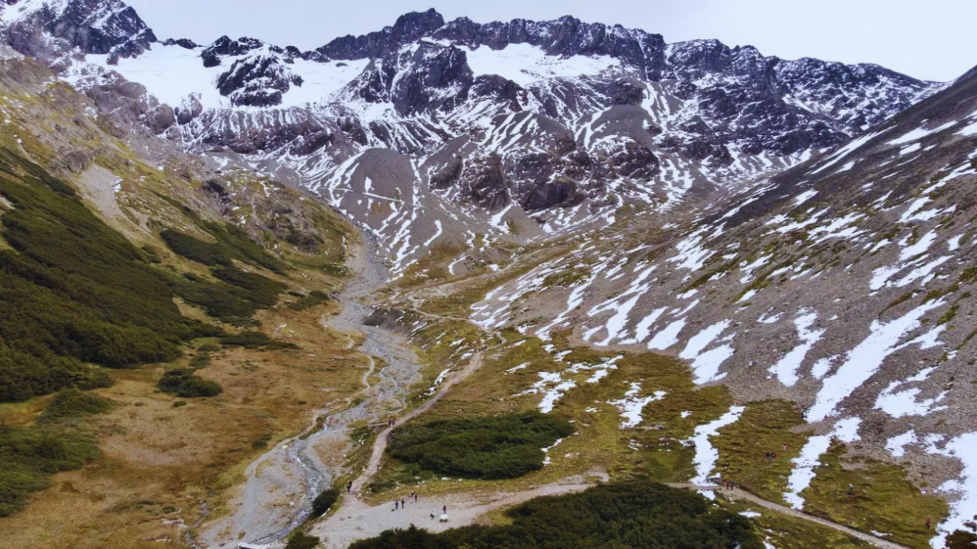 A majestic mountain landscape with patchy snowcaps, rugged terrain, and a meandering river. Green vegetation contrasts with the rocky slopes, creating a serene, inviting ambiance.