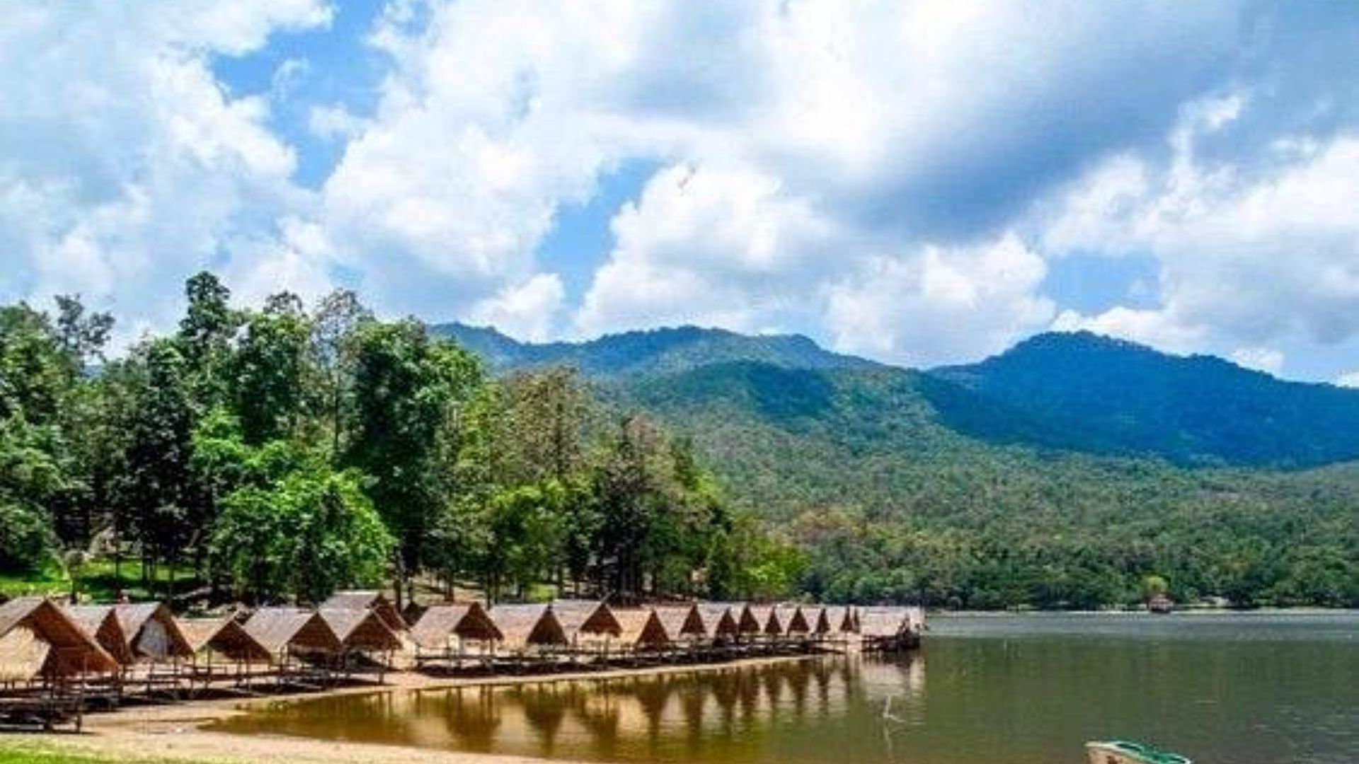 A serene lake bordered by huts, with majestic mountains rising in the background under a clear blue sky.