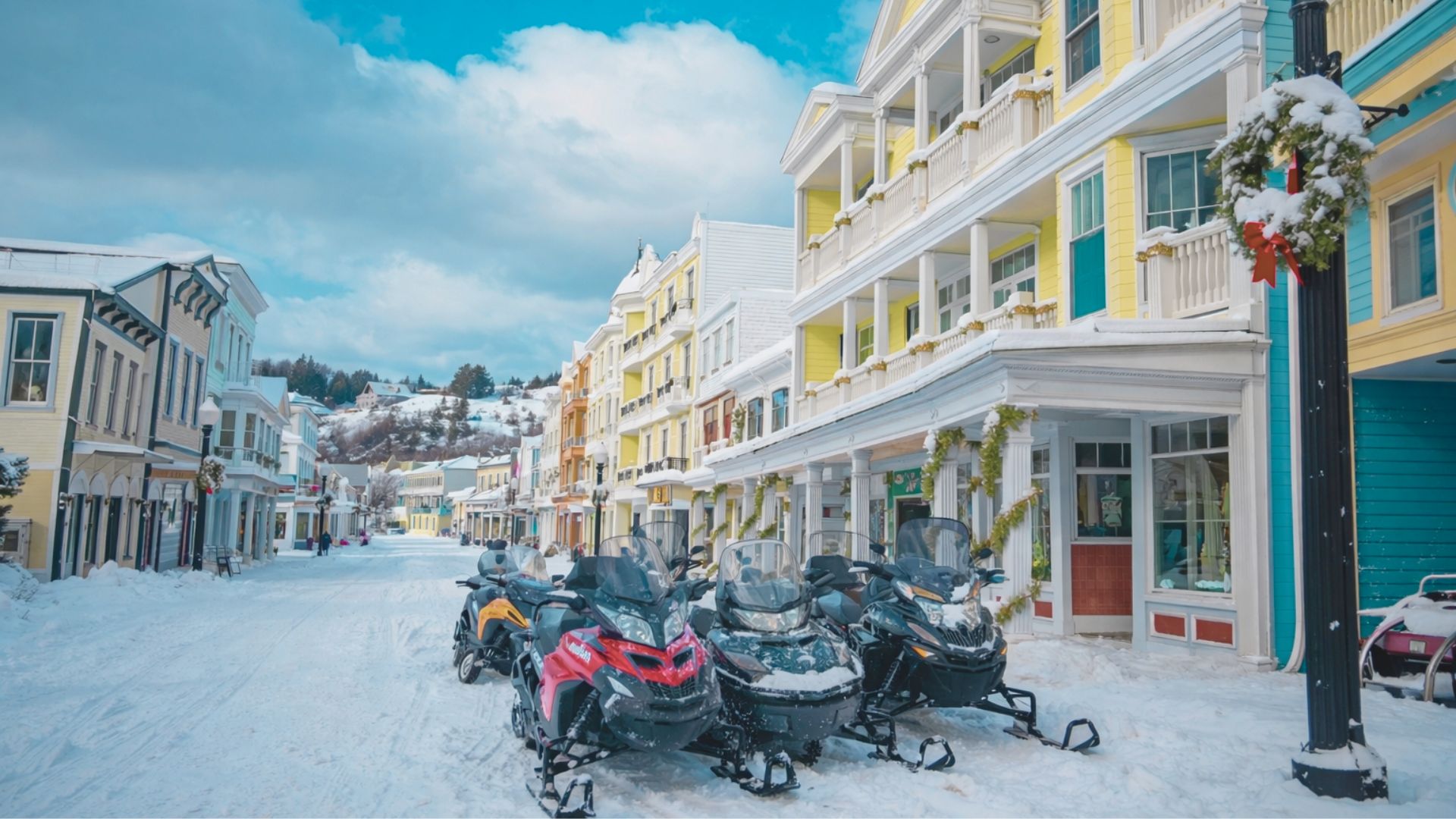 Snowy street lined with colorful buildings and snowmobiles parked in the foreground. The scene depicts a vibrant winter day with a clear blue sky.